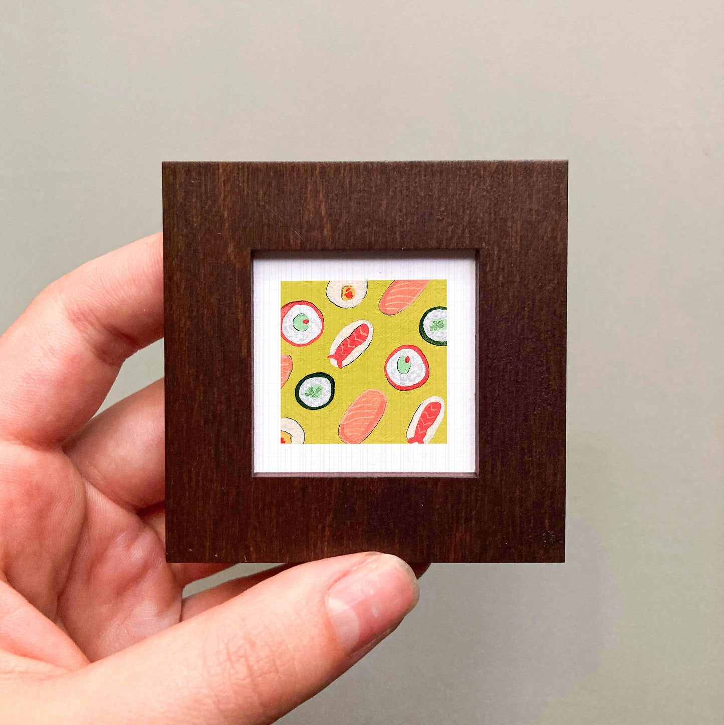A hand is holding a small square picture frame containing a colorful illustration of sushi.