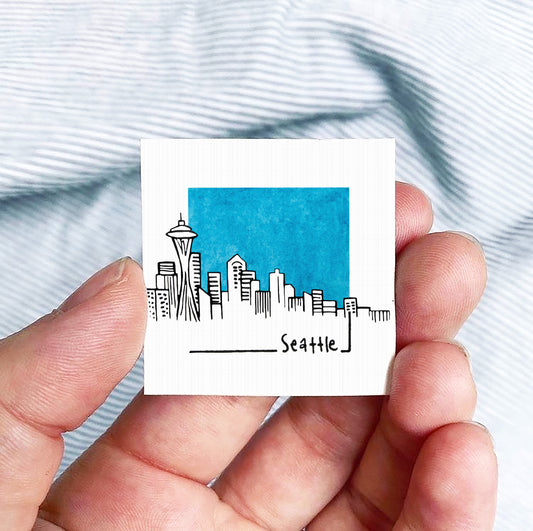 A hand is holding a small square-shaped object with a drawing of the Seattle skyline and the word "Seattle" written on it.