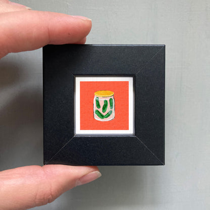 A hand is holding a small black frame containing a framed illustration of a jar of pickles.