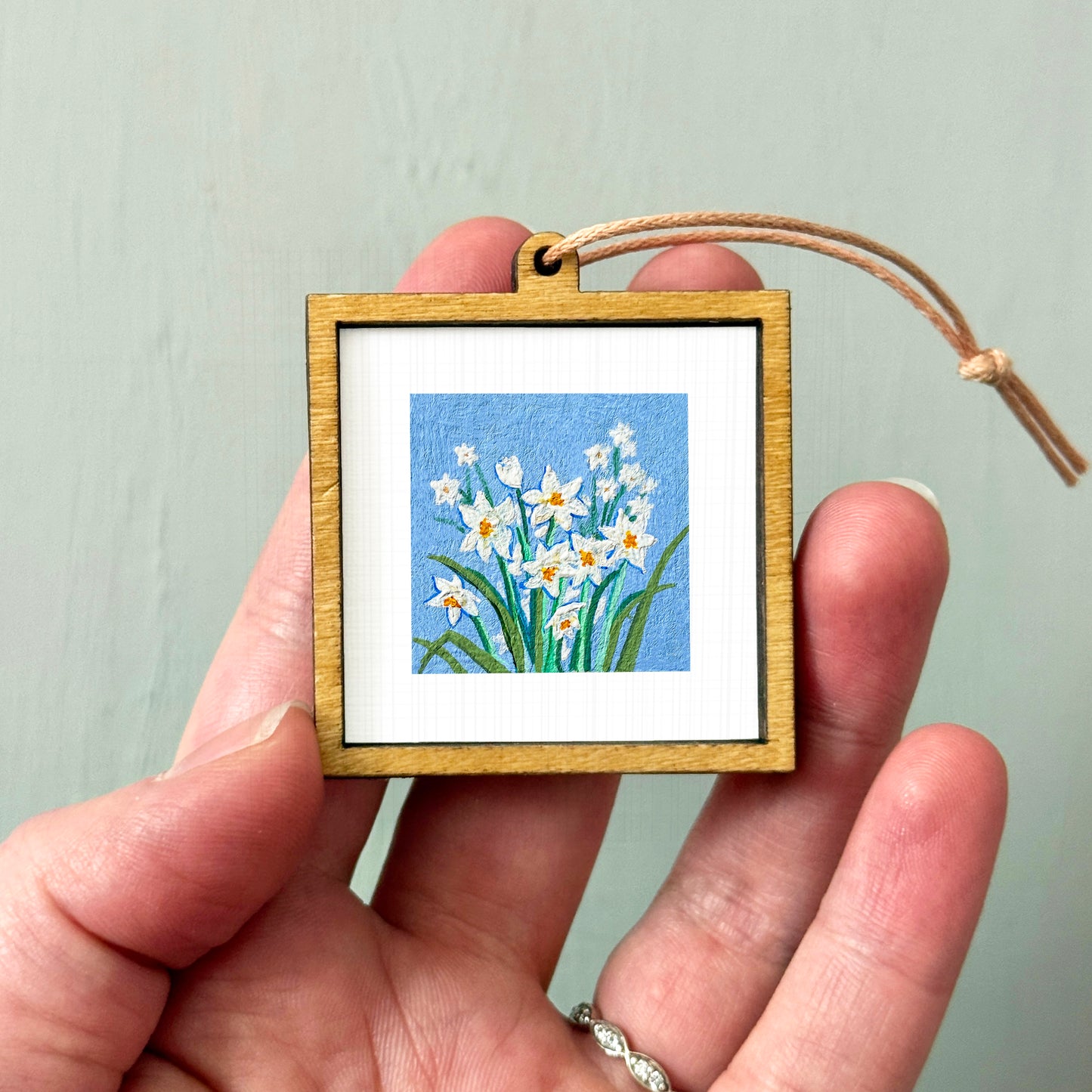 A hand holding a small wooden frame containing a painting of white flowers against a blue background.