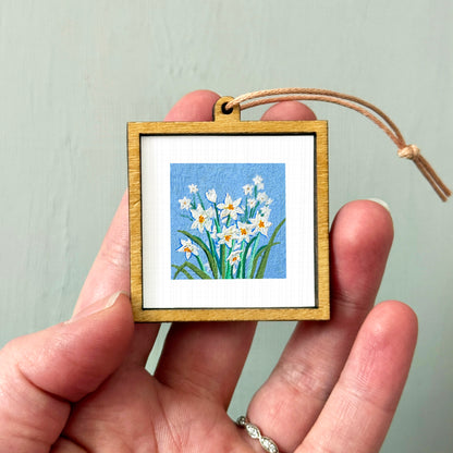 A hand holding a small wooden frame containing a painting of white flowers against a blue background.