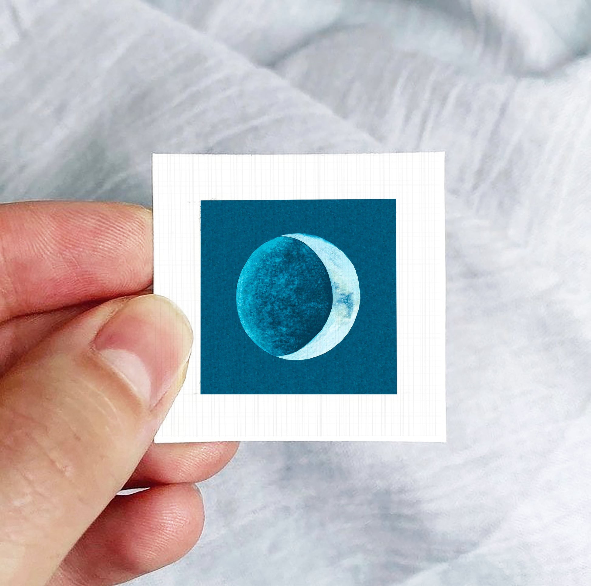 A hand is holding a small square-shaped paper with a blue and white image of a crescent moon on it.