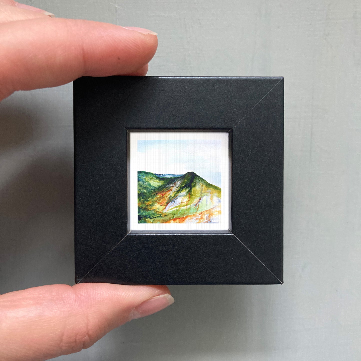 A person's hand holding a small black frame containing a framed watercolor painting of a mountain landscape.