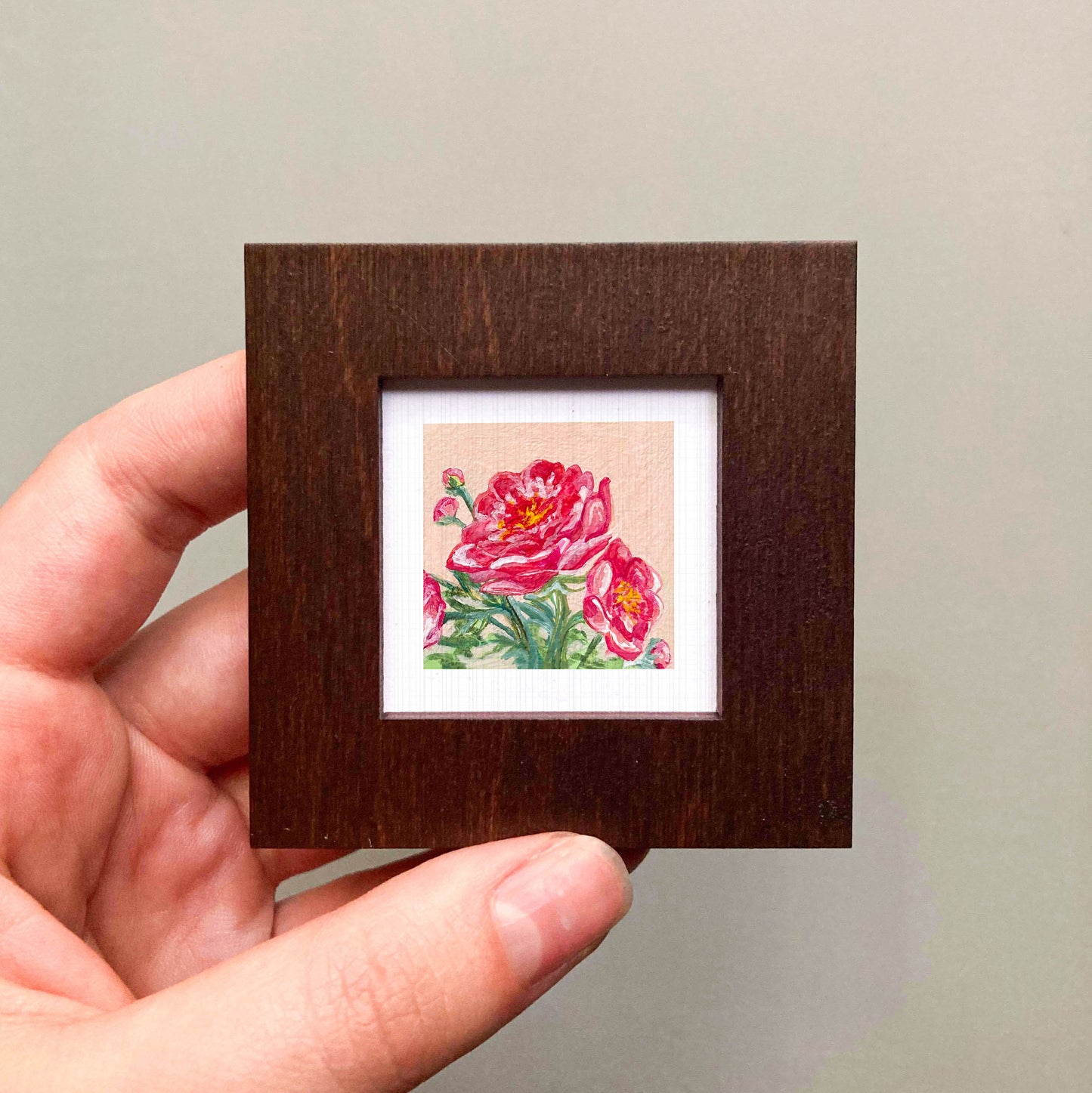 A hand is holding a small wooden frame containing a framed painting of a vibrant red flower.