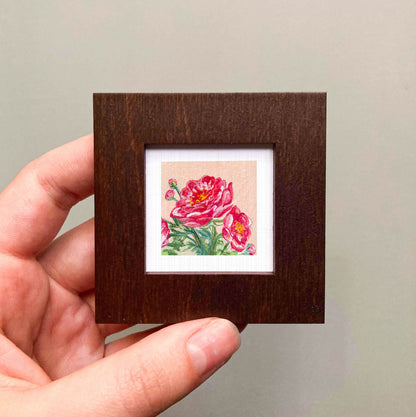 A hand is holding a small wooden frame containing a framed painting of a vibrant red flower.