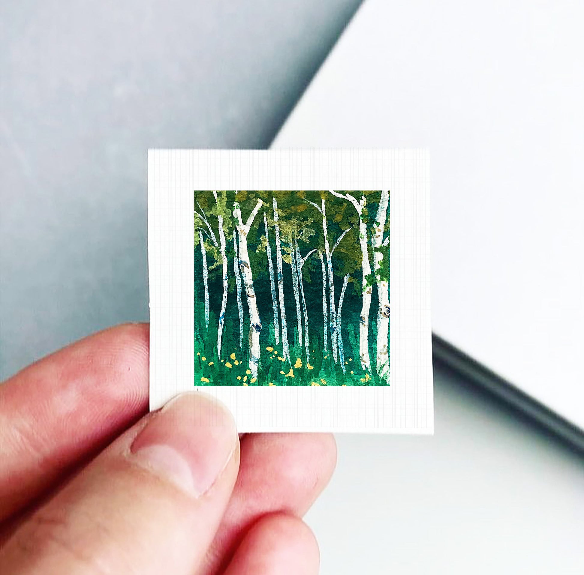 A hand holding a small square painting depicting a forest scene with tall trees and green grass.