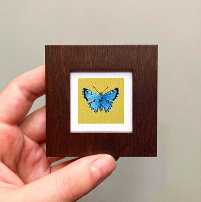 A hand is holding a small wooden frame containing a framed image of a blue butterfly.