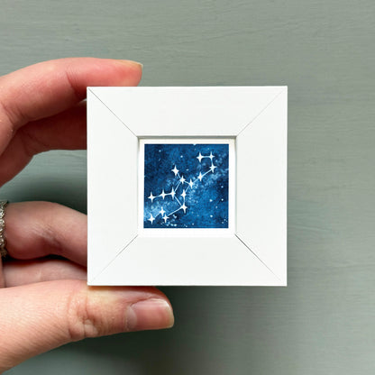 A hand is holding a small white frame containing a painting of a starry night sky.
