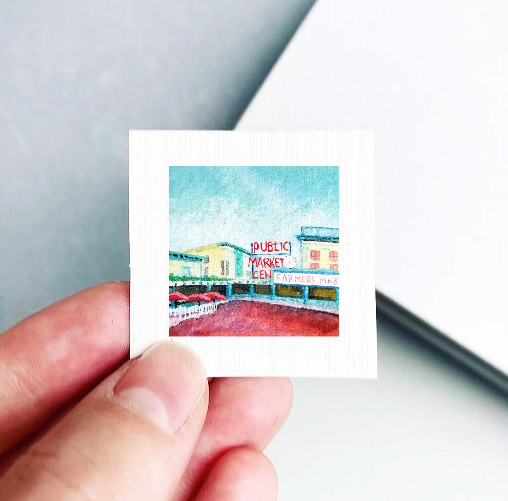 A hand holding a small square picture depicting a colorful scene with a sign that reads "Public Market".