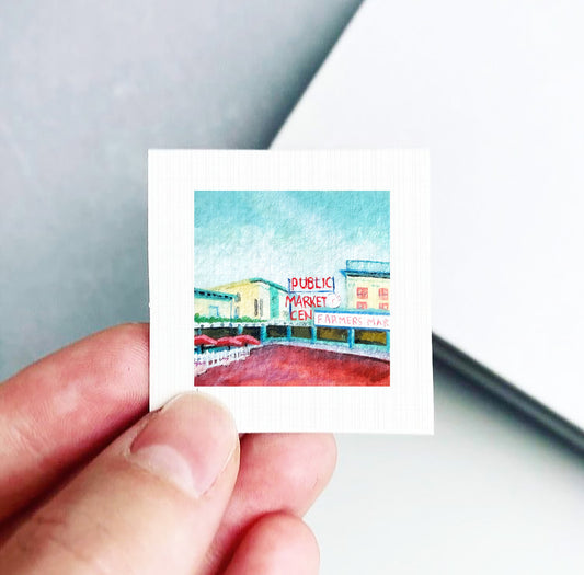 A hand holding a small square picture depicting a colorful scene with a sign that reads "Public Market".