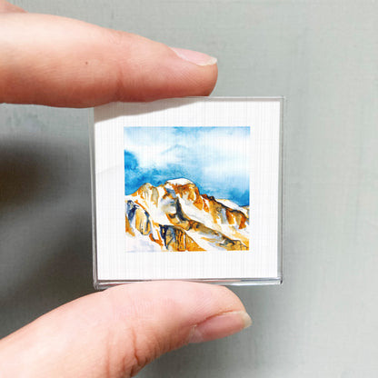 A person's hand holding a small square picture of a mountain landscape with a blue sky and clouds.