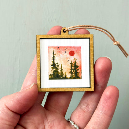 A hand holding a small wooden frame containing a framed painting of a forest scene with a red sun in the sky.