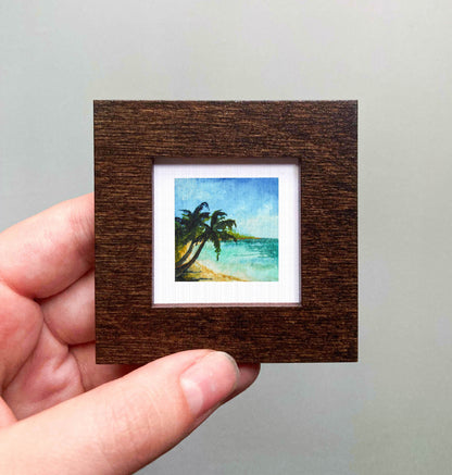 A hand is holding a small, square-shaped picture frame containing a painting of a tropical beach scene with palm trees and a blue sky.