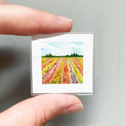 A person's hand is holding a small square picture of a colorful field of flowers.