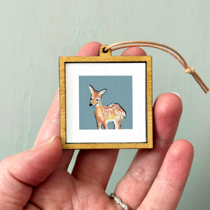 A hand holding a small wooden frame containing a picture of a deer.