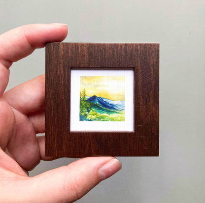 A hand is holding a small wooden frame containing a watercolor painting of a mountain landscape.