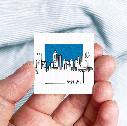 A hand holding a small square-shaped object with a drawing of a city skyline and the word "Atlanta" written on it.