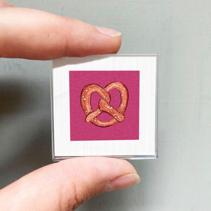 A person's hand holding a small square with a pink background and a heart-shaped illustration of a pretzel.