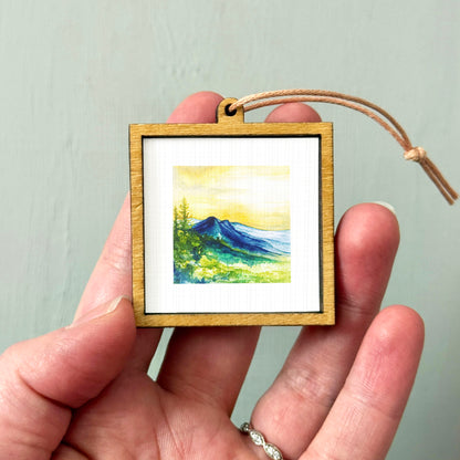 A hand holding a small wooden frame containing a watercolor painting of a mountain landscape.