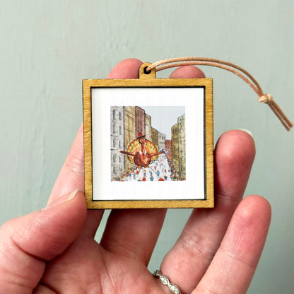 A person's hand is holding a small wooden frame containing a framed illustration of a person walking down a city street.