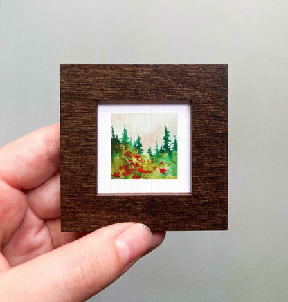 A hand is holding a small, square-shaped wooden frame containing a watercolor painting of a forest scene with pine trees and red flowers.