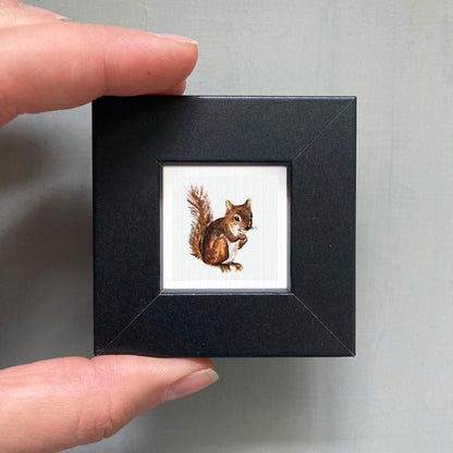 A person's hand holding a small black square frame containing a painting of a squirrel.