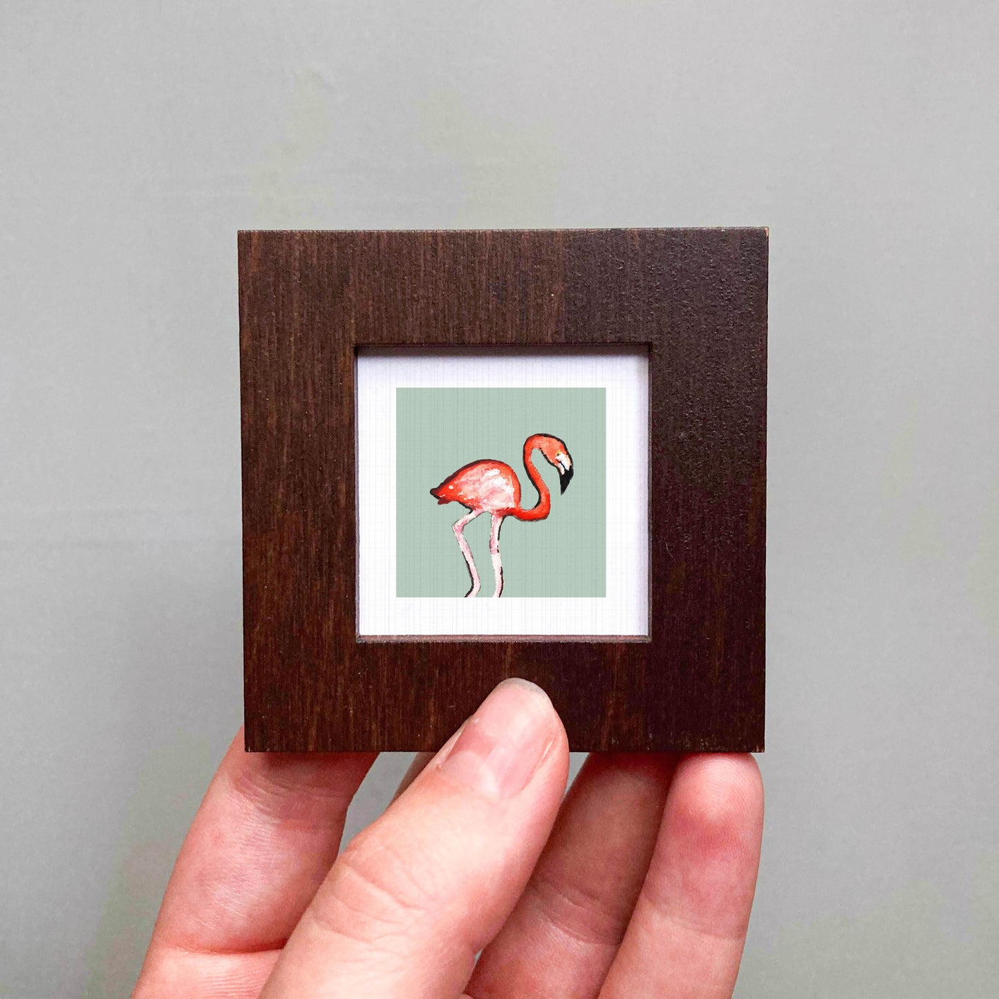A person's hand is holding a small wooden frame containing a framed illustration of a flamingo.