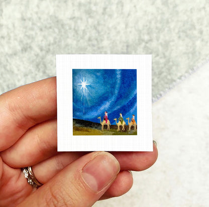 A hand holding a small square picture depicting a scene with three people and a star in the sky.