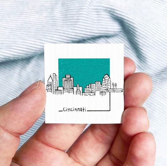 A hand holding a small square-shaped object with a drawing of a city skyline on it, against a striped background.
