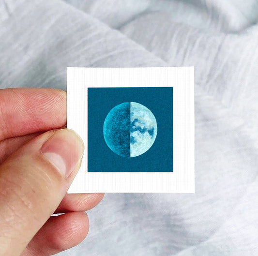 A hand is holding a small square-shaped object with a blue and white image of a moon on it.