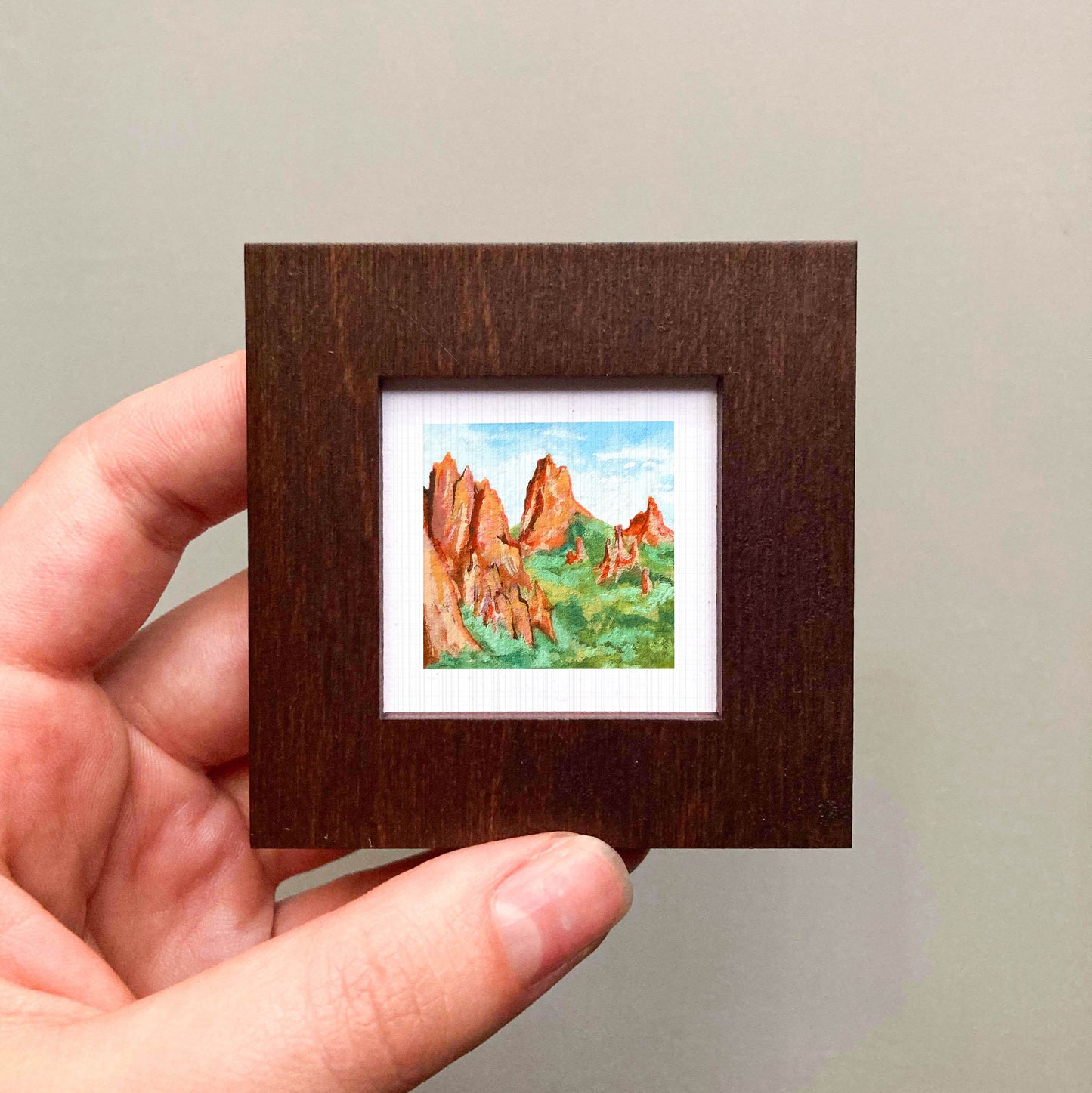 A hand is holding a small, square-shaped wooden frame containing a watercolor painting of a mountain landscape.