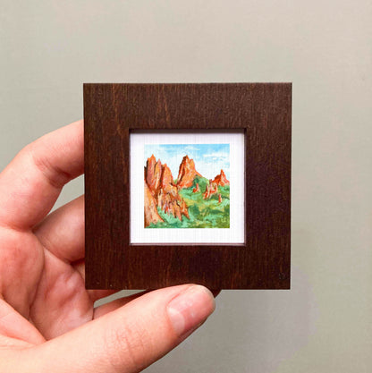 A hand is holding a small, square-shaped wooden frame containing a watercolor painting of a mountain landscape.