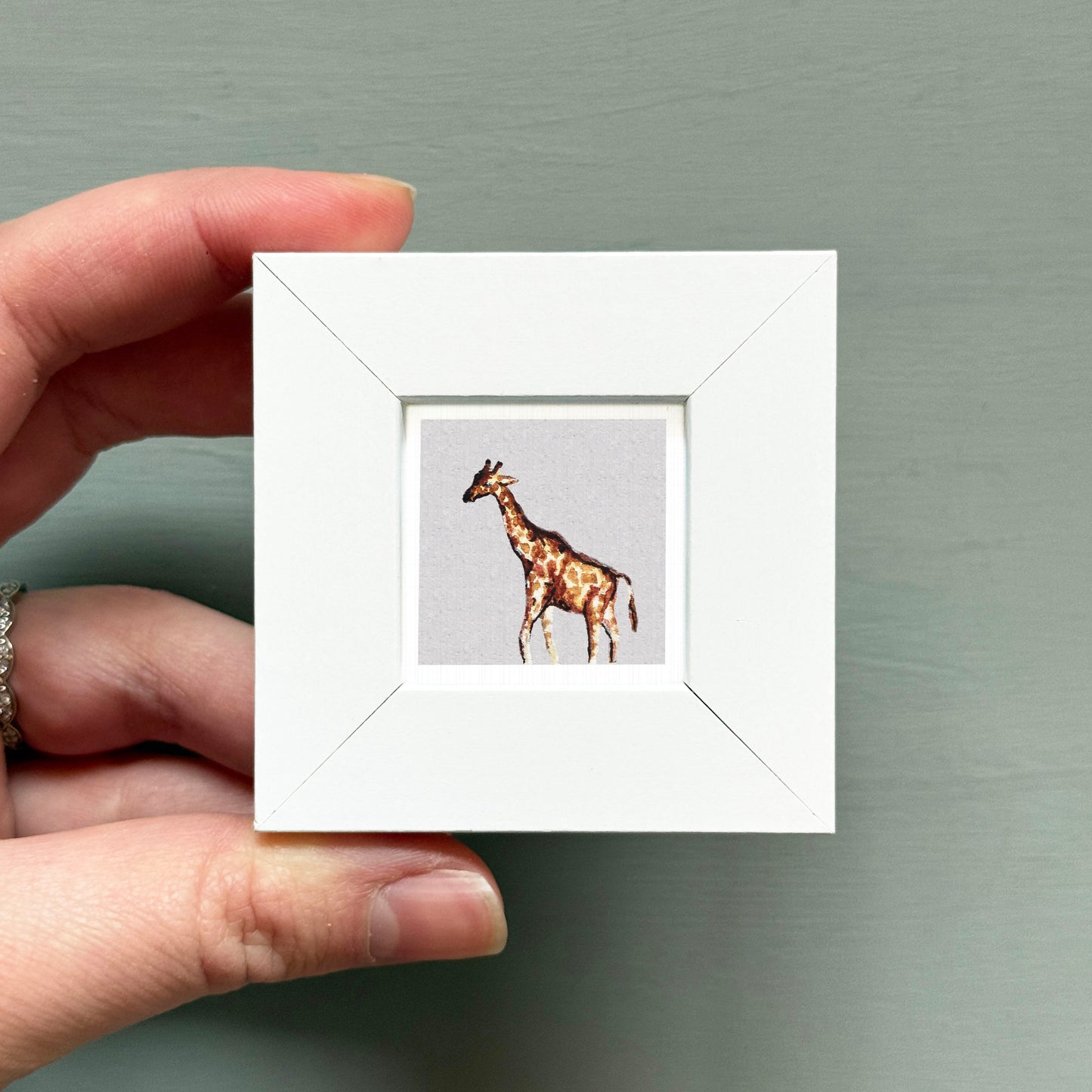 A hand is holding a small white frame containing a picture of a giraffe.