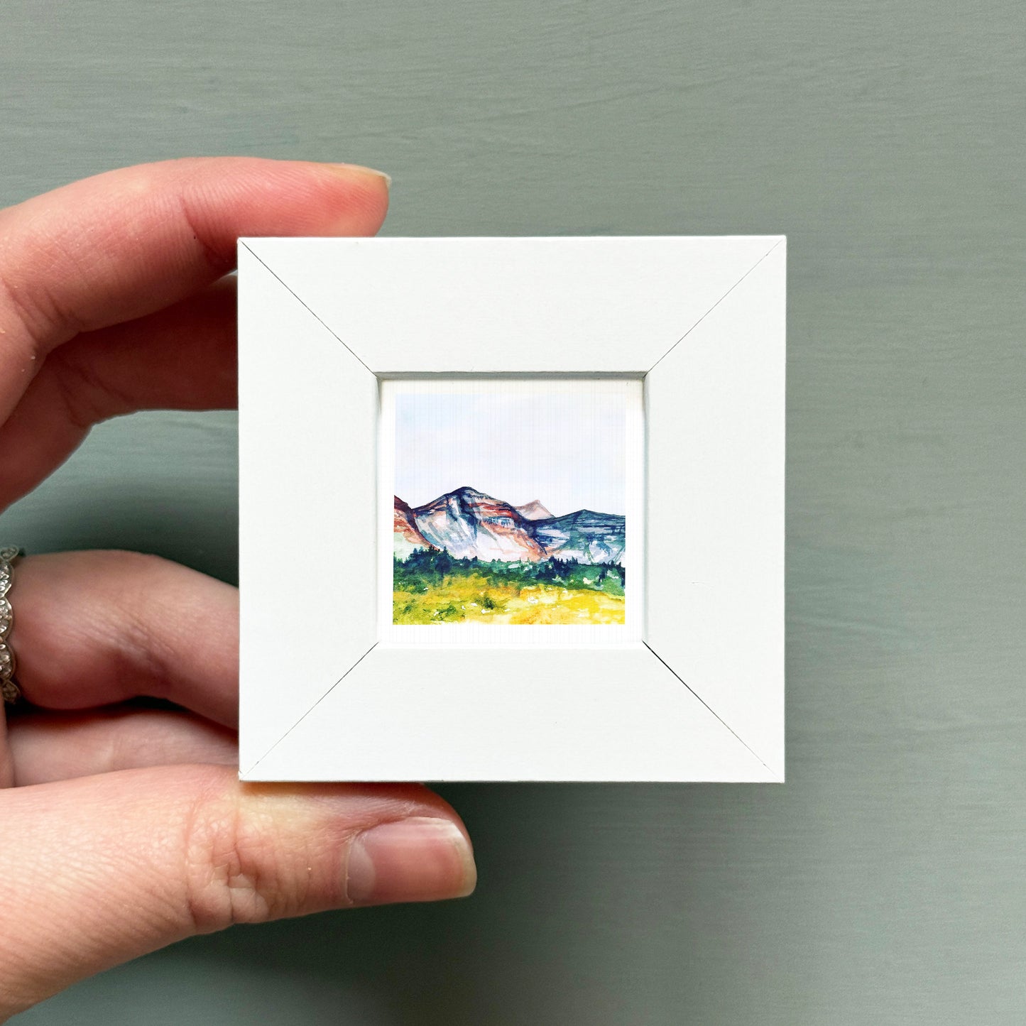 A hand is holding a small white square frame containing a watercolor painting of a mountain landscape.