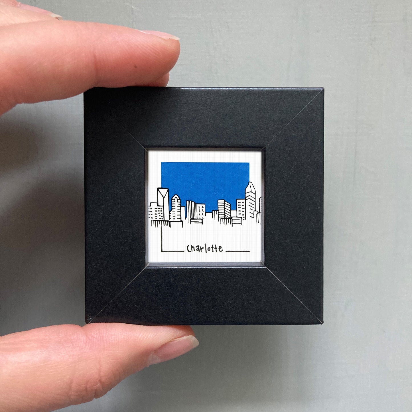 A person's hand is holding a small black square frame containing a framed illustration of a city skyline with a blue sky.