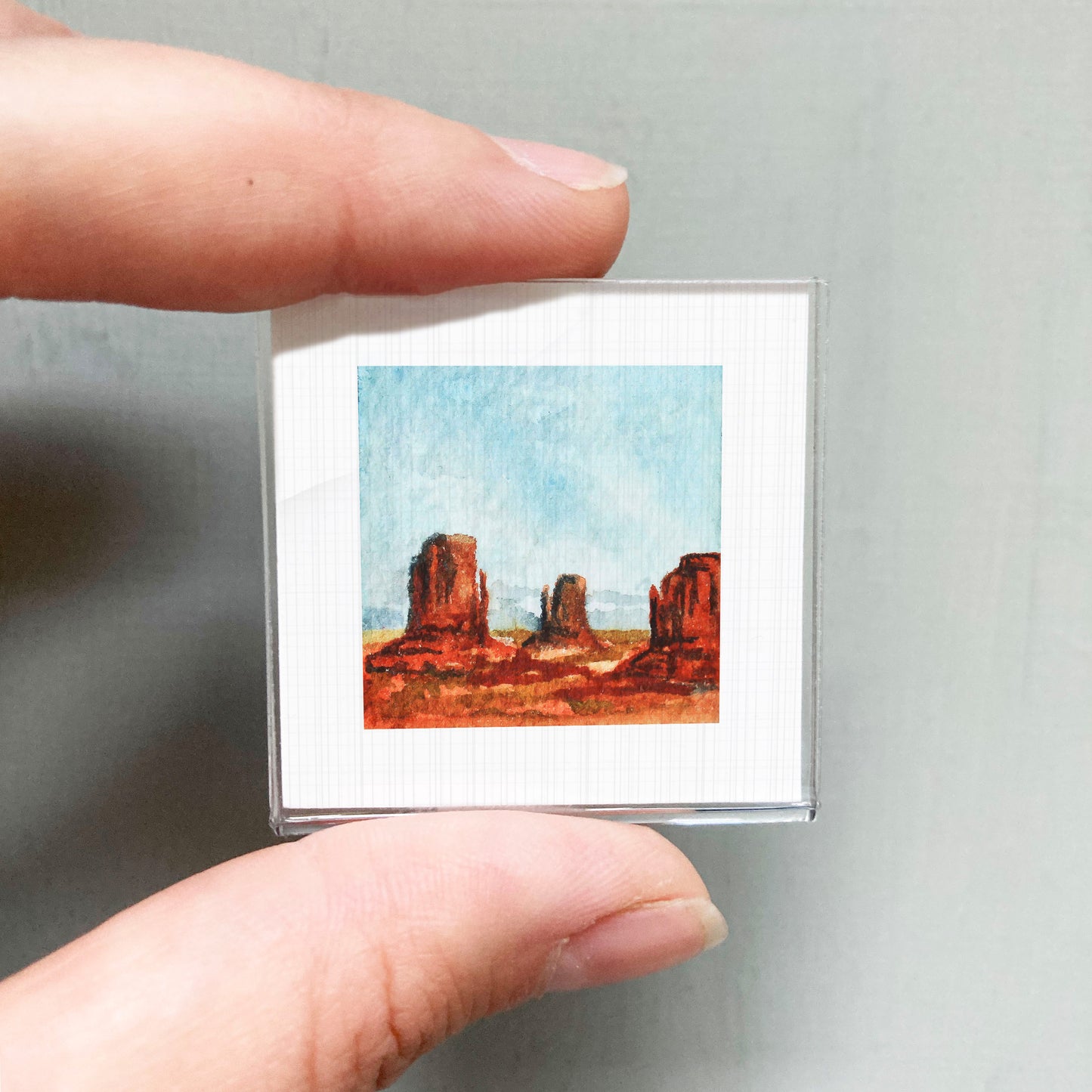 A person's hand holding a small square picture of a desert landscape with red rock formations.