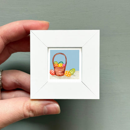 A hand is holding a small white picture frame containing an illustration of a basket filled with colorful Easter eggs.