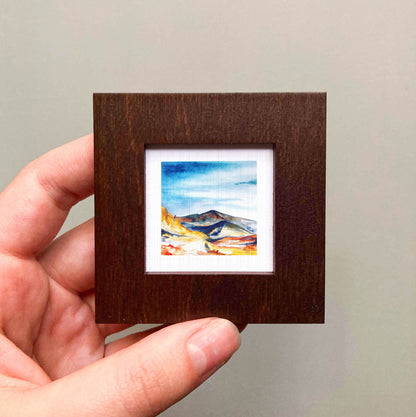 A hand is holding a small, square-shaped wooden frame containing a watercolor painting of a mountain landscape.
