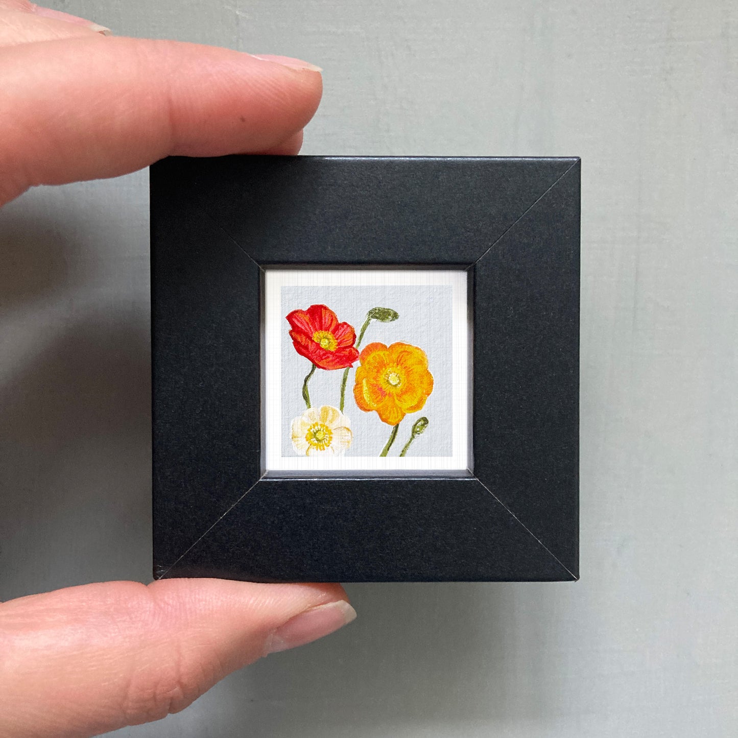 A person's hand holding a small black frame containing a framed painting of two flowers, one red and one yellow.