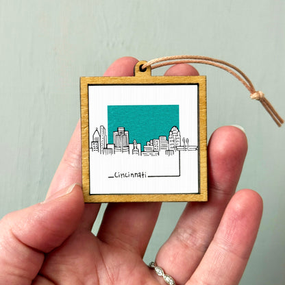 A hand holding a small wooden frame with a picture of the Cincinnati skyline inside it.