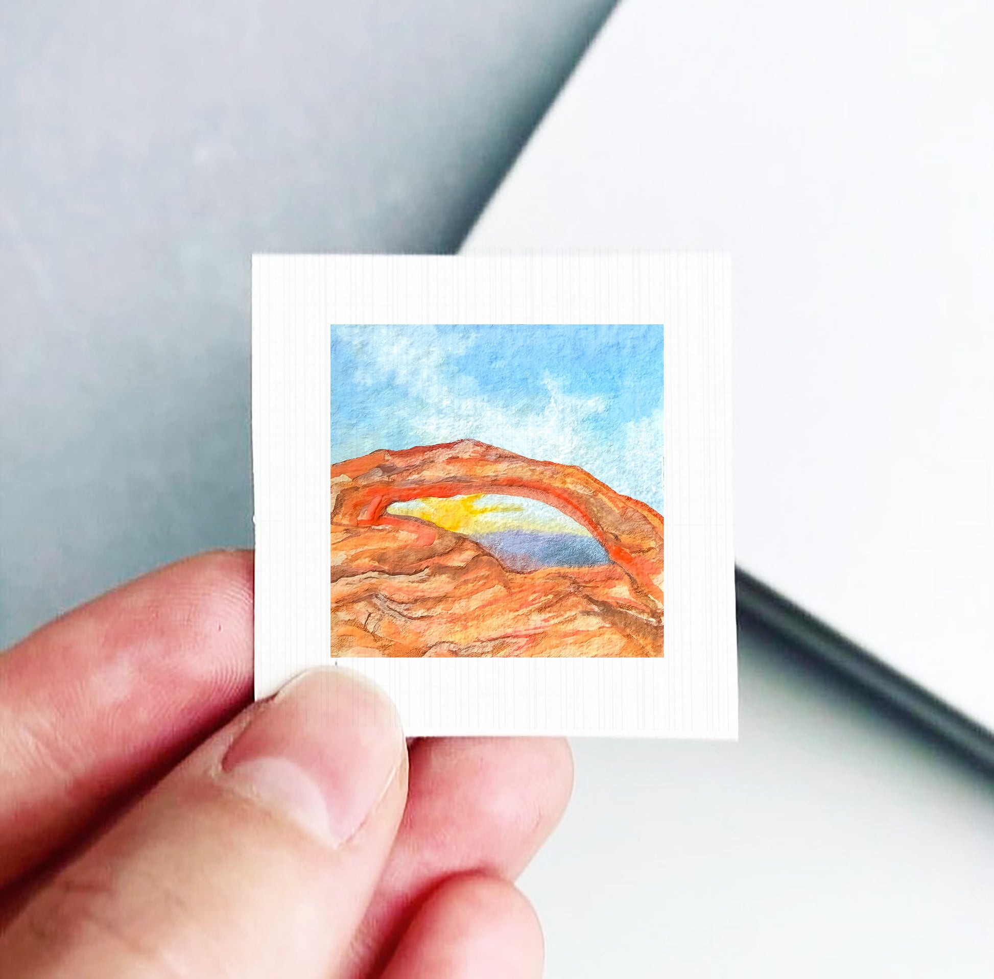 A hand is holding a small square piece of paper with a watercolor painting of a desert landscape on it.