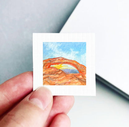 A hand is holding a small square piece of paper with a watercolor painting of a desert landscape on it.