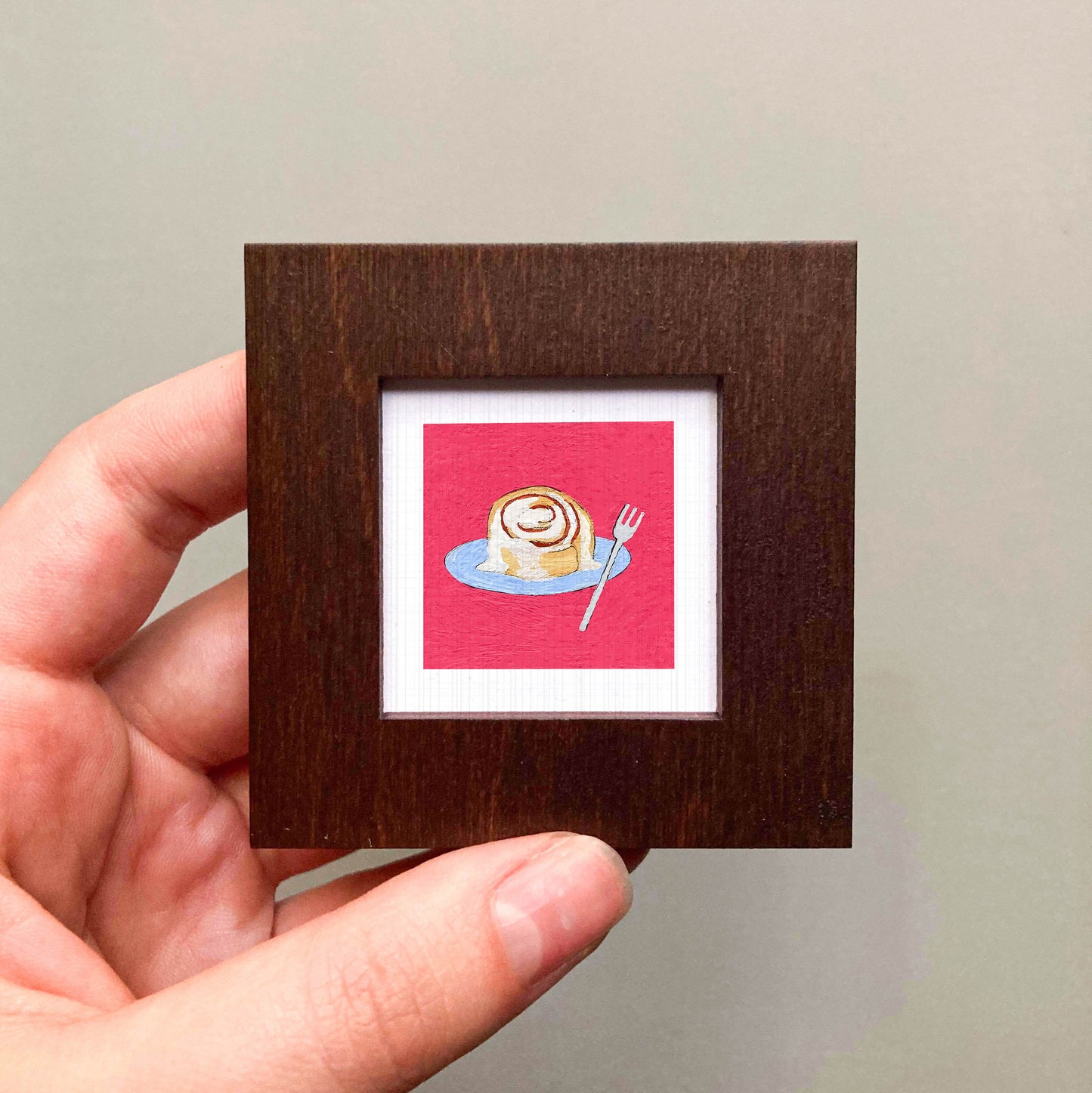 A hand is holding a small wooden frame containing a framed illustration of a cinnamon roll on a plate.