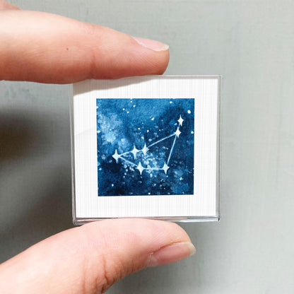 A hand is holding a small square-shaped object with a blue background and white stars, which appears to be a piece of art or a decorative item.