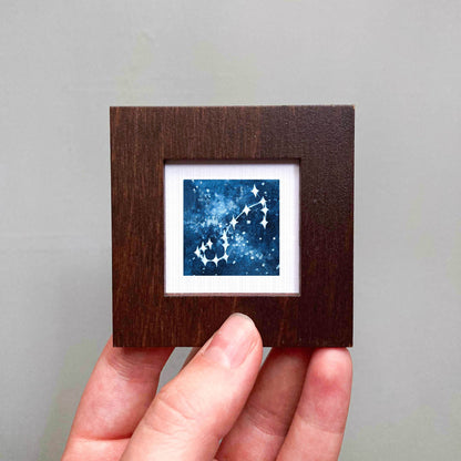 A hand is holding a small wooden frame containing a framed artwork depicting a starry night sky.