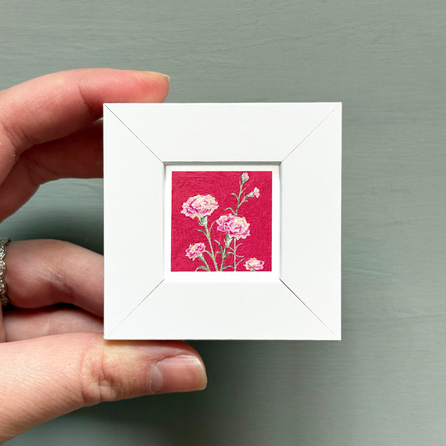 A hand is holding a small white square frame containing a painting of pink flowers against a red background.