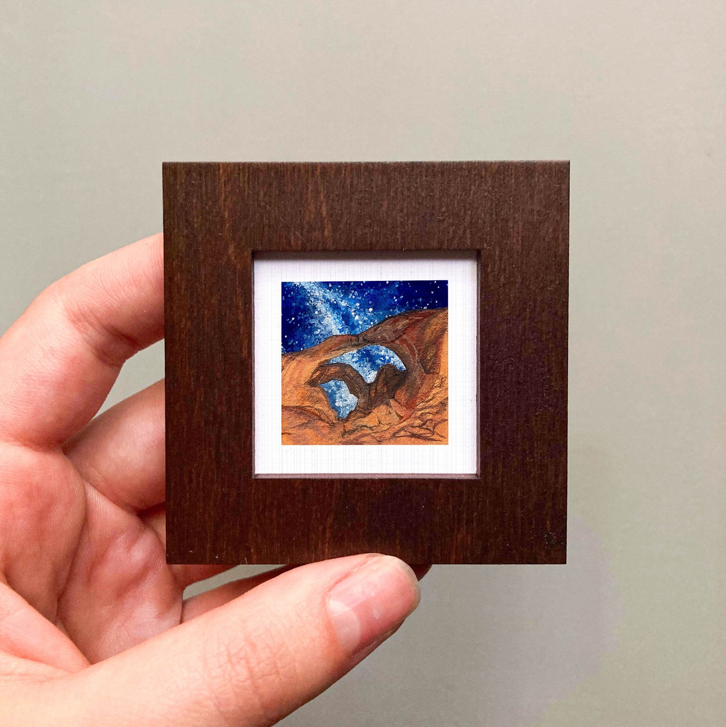 A hand is holding a small wooden frame containing a painting of a desert landscape with a starry night sky.