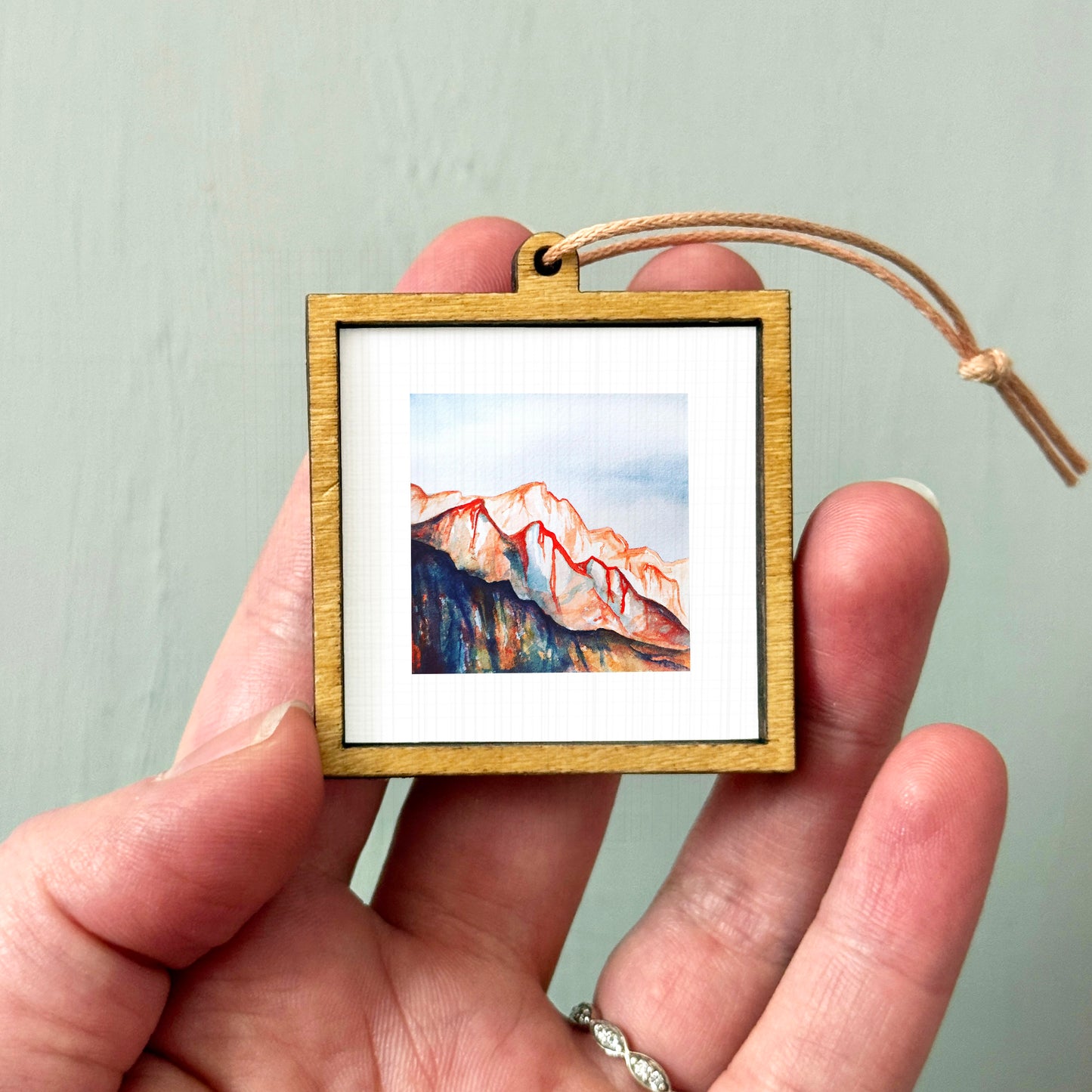 A hand holding a small wooden frame containing a watercolor painting of a mountain landscape.