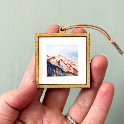 A hand holding a small wooden frame containing a watercolor painting of a mountain landscape.