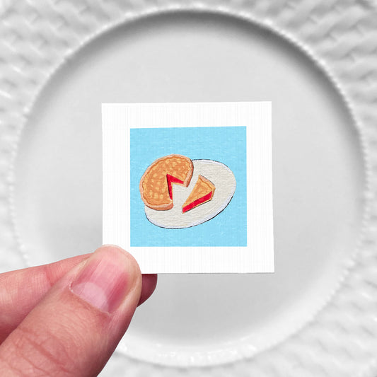 A hand is holding a small square card with an embroidered image of a piece of food, specifically a waffle or pancake, on a blue background.